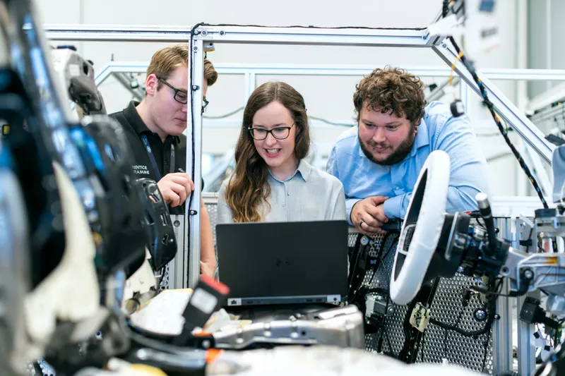 Three people - 2 man and 1 woman looking at laptop in manufacturing environment.